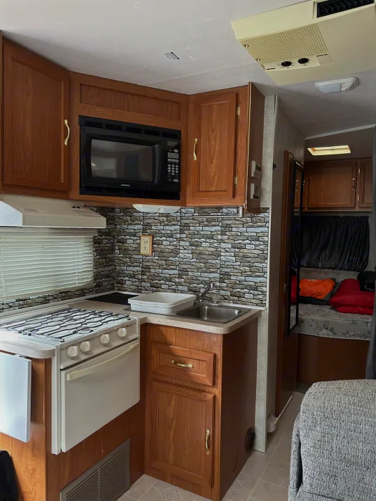 Inside an RV rental: kitchen area with stove, sink, microwave, and wood cabinets. Bed visible in background.. Coachmen Catalina 2003