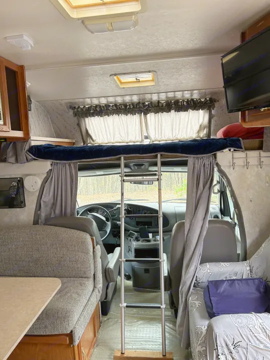 An RV rental interior shows the over-cab bunk with ladder, front cabin, and dinette area, parked with a view of trees.. Coachmen Catalina 2003