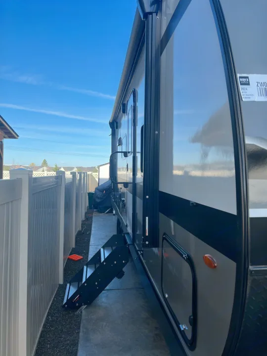 Side view of a 2024 Jayco Grey Feather travel trailer, ideal for your next RV rental adventure. Steps extend, parked by a fence.. Jayco Jayflight 2024