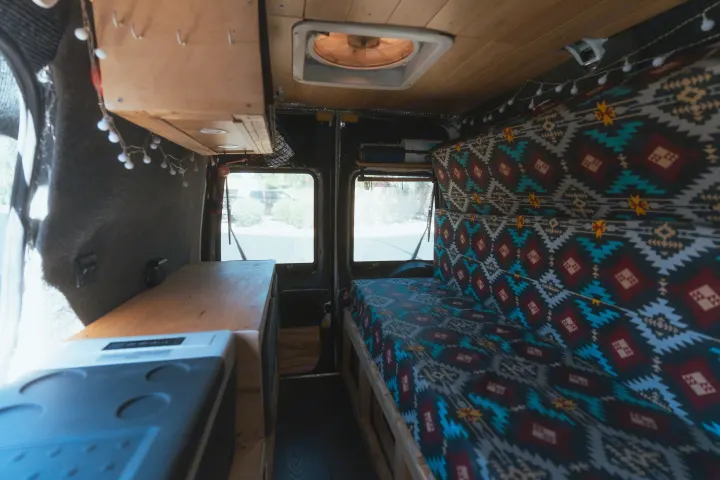 Cozy RV rental camper van interior. Features a colorful patterned bed, wooden cabinets, and a portable fridge.. Ford Transit 2011