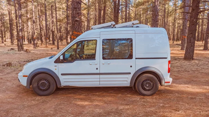 White Ford Transit Connect camper van, ideal for rv rental, parked in a sunlit pine forest. Trees have orange markings.. Ford Transit 2011