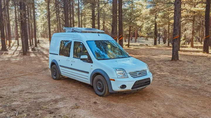 White Ford Transit Connect camper van with solar panels, perfect as an RV rental, parked in a sunlit pine forest.. Ford Transit 2011