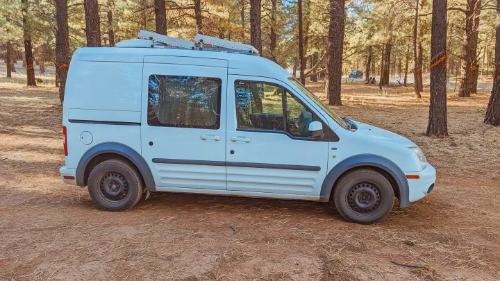 White Ford Transit Connect camper van with roof rack, parked in a sunny pine forest. Perfect RV rental for adventure.. Ford Transit 2011