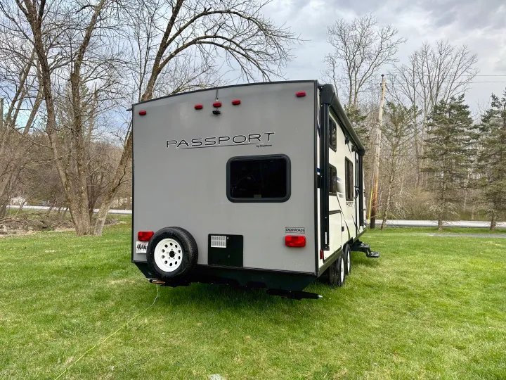 Grey RV rental travel trailer parked on green grass with bare trees. Ideal for outdoor adventures.. Keystone RV Passport Express 2019
