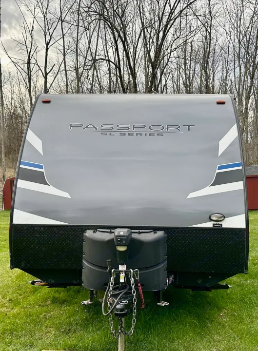 Front view of a gray "Passport SL Series" travel trailer, perfect for an RV rental getaway, parked on green grass under bare trees.. Keystone RV Passport Express 2019