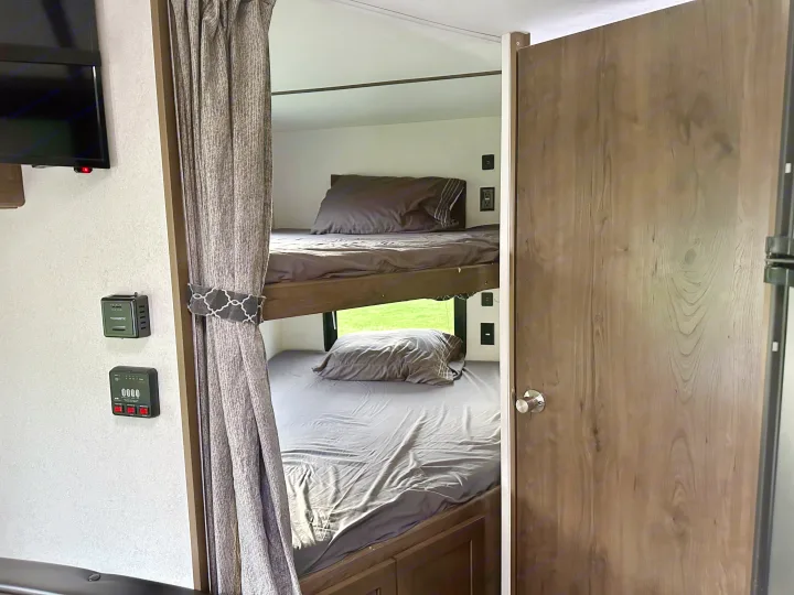 Interior view of an **RV rental** showing modern bunk beds with grey bedding, a privacy curtain, and control panels.. Keystone RV Passport Express 2019
