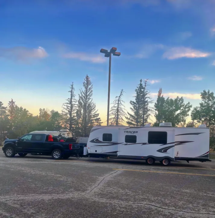 Dark pickup truck towing a white Tracer RV rental in a parking lot. Gear visible in truck bed, trees under blue sky.. Forest River Tracer Executive 2010