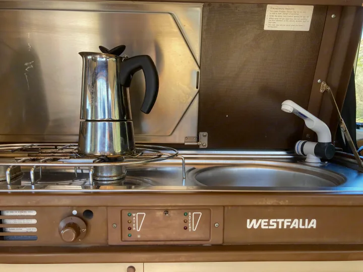 An RV rental's compact Westfalia kitchen with a Moka pot on the stove, sink, and water/battery level indicators.. Volkswagen Westfalia 1986