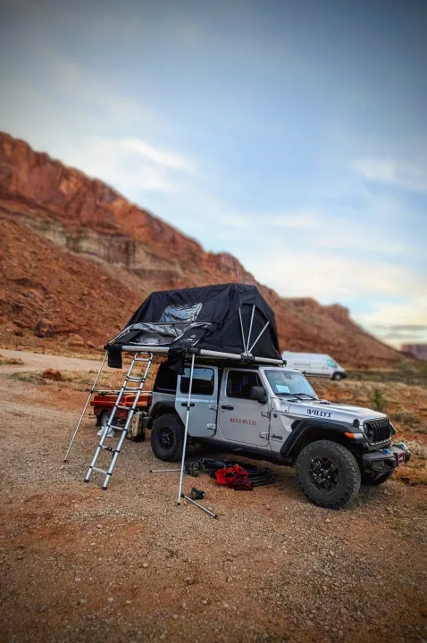 An adventure-ready RV rental Jeep with a rooftop tent set up in a vast desert landscape with red rock formations.. Jeep Wrangler 2024