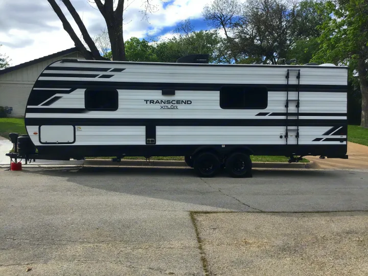 White Transcend Xplor travel trailer, a potential RV rental, parked on asphalt. Features black graphics, dual axles, & rooftop AC.. Grand Design Transcend Xplor 2024