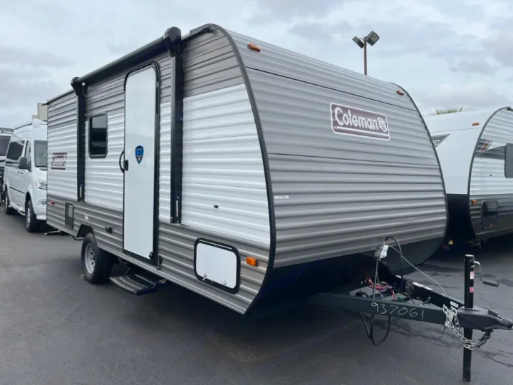 This gray Coleman travel trailer, ideal for an RV rental, is parked in a lot, featuring a side door and an awning.. Coleman Lantern LT 17 B 2024