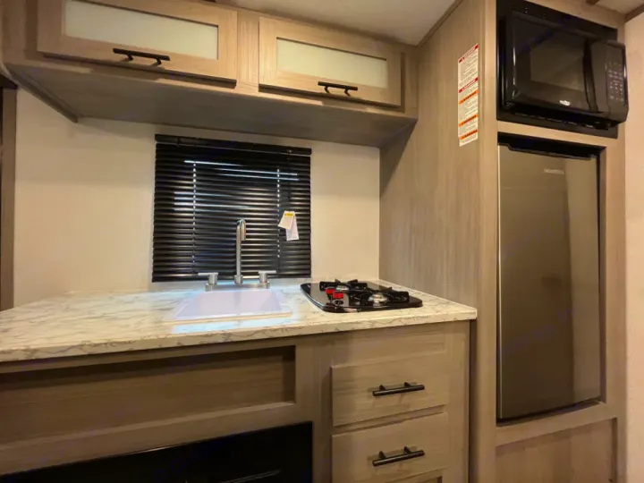 Compact RV rental kitchen with a white sink and two-burner cooktop on a light counter, next to a microwave and fridge.. Coleman Lantern LT 17 B 2024