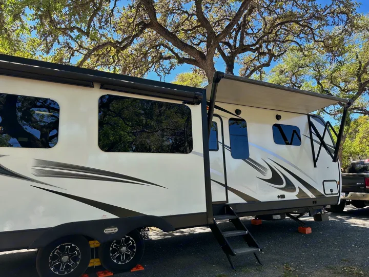 White RV rental with awning extended, stabilized on orange blocks, parked under sunny oak trees at a campground.. Venture STT343VIB 2022