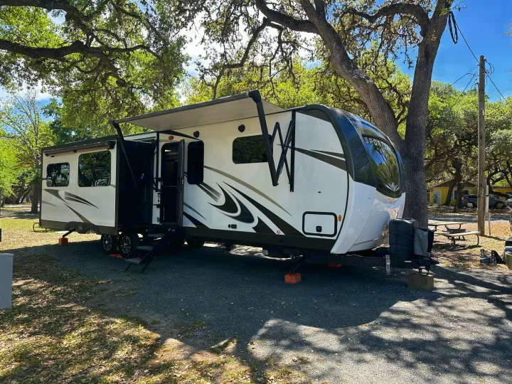 White RV rental travel trailer with awning extended, set up on a gravel site under large shade trees. Ideal for camping.. Venture STT343VIB 2022