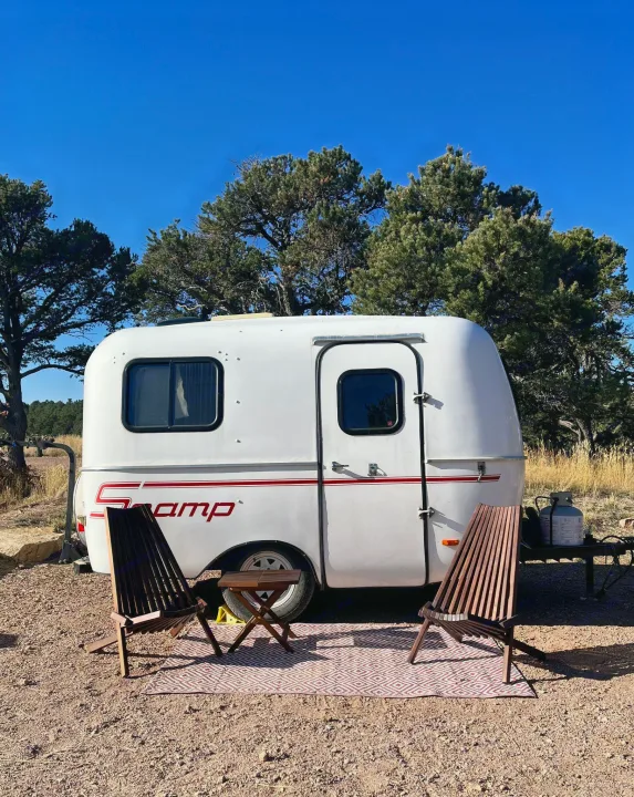 This cozy Scamp travel trailer setup, perfect for an rv rental, features chairs & a table on a rug at a scenic campsite under a blue sky.. Scamp Scamp Trailer 2004