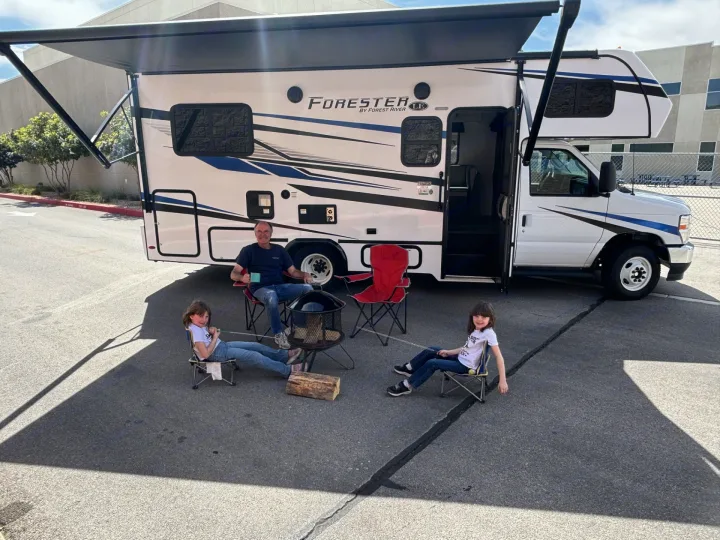 RV rental family fun! Dad and two kids relax by a fire pit under their motorhome's extended awning.. Forester LE 2024