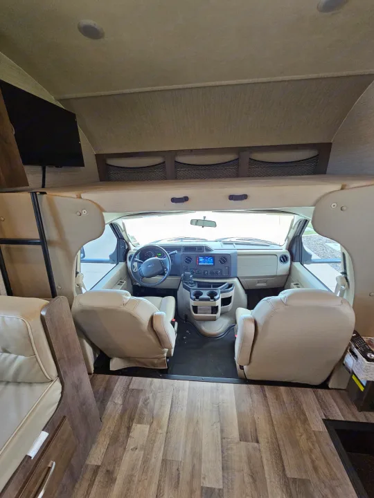 RV rental interior: Spacious front cockpit with tan driver/passenger seats, dashboard, and overhead bunk.. Jayco Redhawk 2020