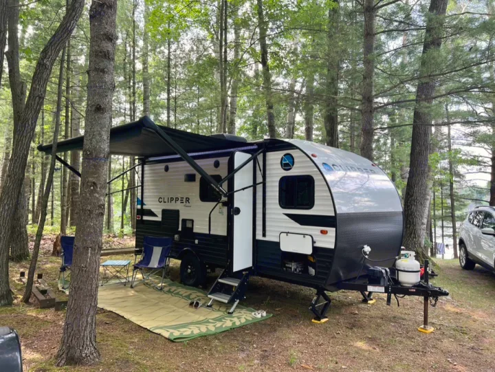 This rv rental, a white and black travel trailer, is set up at a serene wooded lake campsite with an extended awning, chairs, and mat.. Clipper Ultra Lite 2022