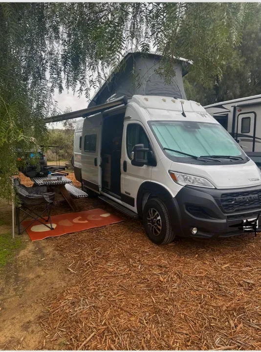 An RV rental camper van with an open pop-top roof and extended awning, set up at a wooded campsite with a picnic area.. Winnebago Solis 2023
