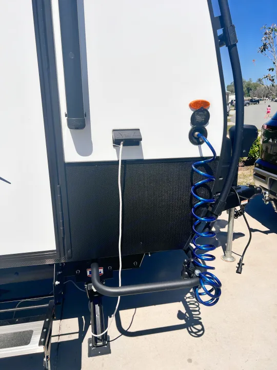 Side view of an RV rental at a campground. Power cable & blue coiled water hose are connected for setup.. Winnebago Hike 100 2023