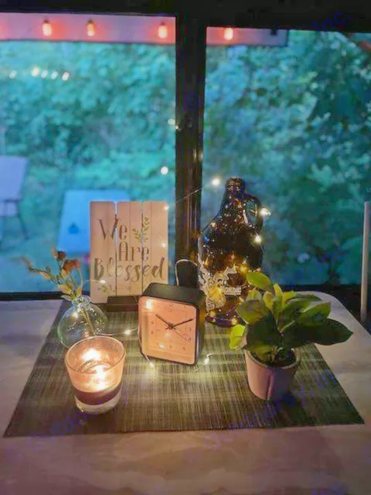 Relaxing RV rental evening setup: lit candle, string lights, clock, and plant on table, with the forest outside.. Forest River Wildwood X-Lite 2020