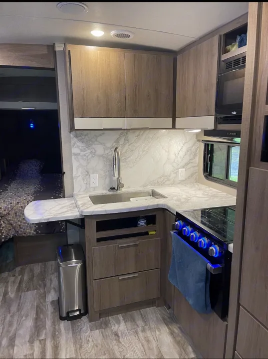 RV rental interior: A compact, modern kitchen with a sink, stove, microwave, and wood-look cabinets. Bedroom visible left.. Grand Design imagine 2020