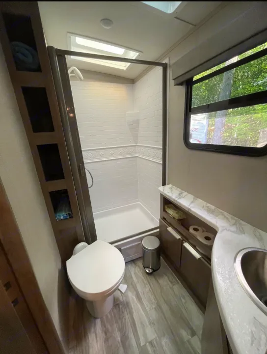 This RV rental bathroom offers a compact shower, toilet, sink vanity, and storage shelves.. Grand Design imagine 2020