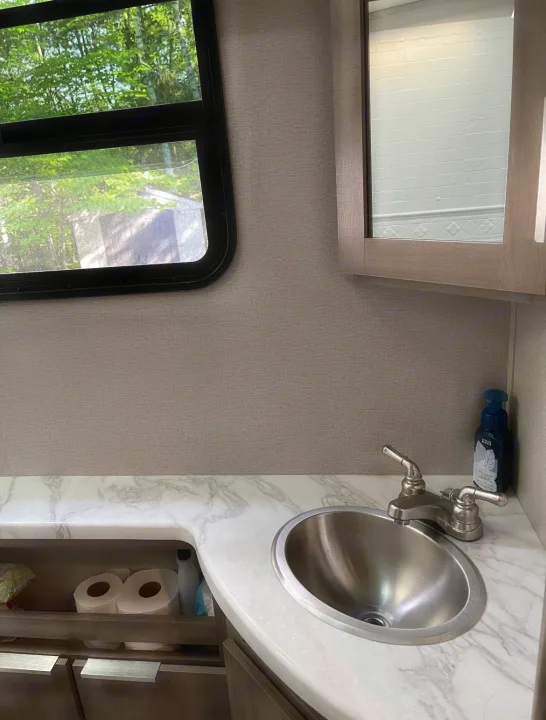 Inside an RV rental: a compact bathroom vanity with a stainless sink, marble-look counter, mirror, and window.. Grand Design imagine 2020