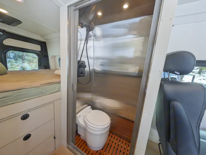 This RV rental interior showcases a compact wet bath with a toilet and shower, a cozy bed with storage, and scenic window views.. New Life Conversions Luxury Edition 2022