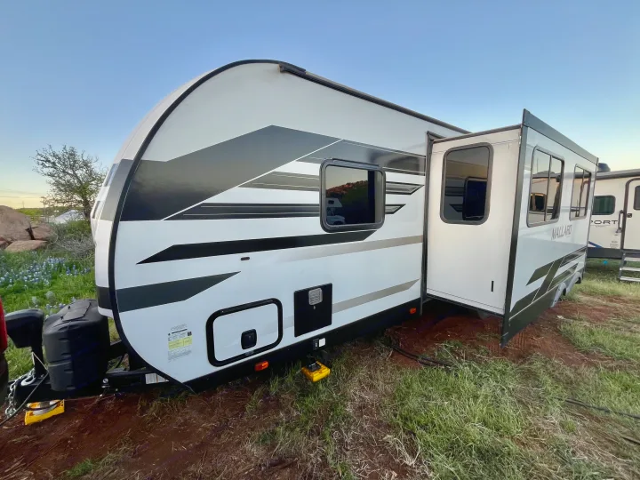 White travel trailer RV rental with extended slide-out, parked on a dirt lot with grass. Perfect for travel & camping adventures.. Heartland M-3385 2021