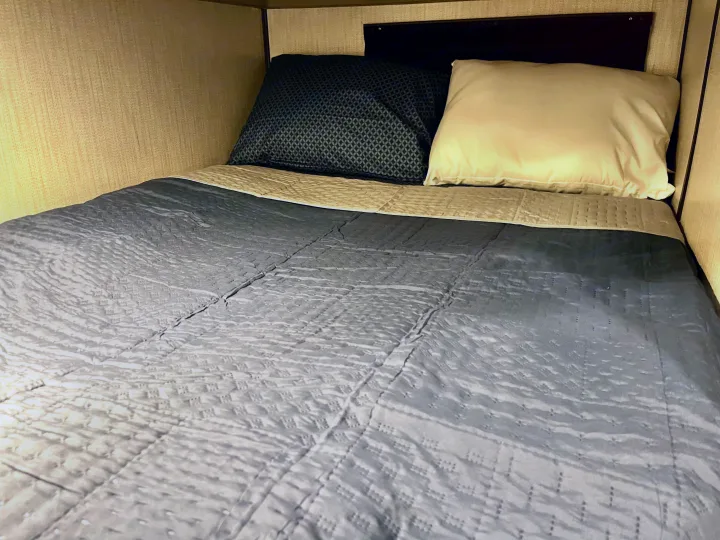 Compact RV rental sleeping area with textured walls, a blue-gray quilted comforter, and patterned and plain pillows.. Heartland M-3385 2021
