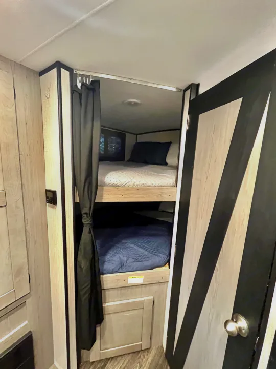 An RV rental's compact sleeping area featuring two bunk beds with blue and white bedding, a privacy curtain, and modern wood-look finishes.. Heartland M-3385 2021