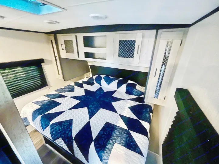 Interior of an RV rental bedroom featuring a bed with a blue and white star quilt, overhead cabinets, and a ceiling skylight.. Heartland M-3385 2021