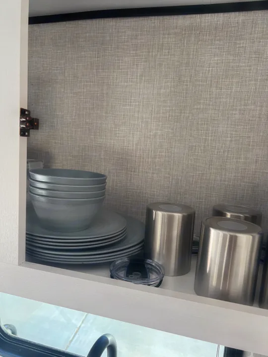Open RV rental kitchen cabinet displaying stackable gray dishes, bowls, and stainless steel travel mugs.. Jayco 224BHW 2024