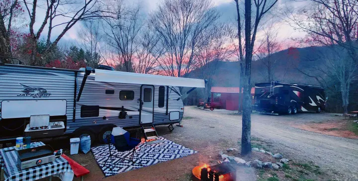 An RV rental travel trailer is set up at a rustic campsite with an outdoor kitchen, rug, campfire, and sunset. A serene mountain view.. Jayco Jay Flight SLX 2021