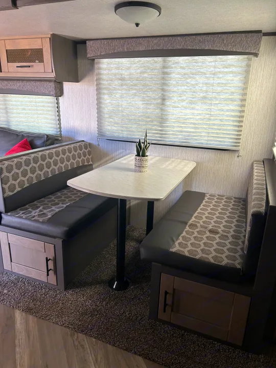 Spacious RV rental dinette with patterned booth seating, a white table, overhead storage, and bright windows.. Shadow Trailers Shadow Cruiser 2020