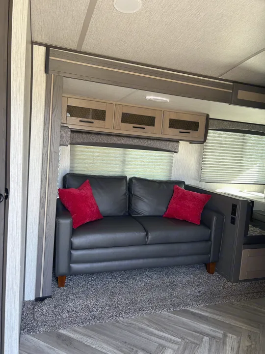 RV rental interior with a dark two-seater sofa, red throw pillows, light wood cabinets, and mixed grey flooring.. Shadow Trailers Shadow Cruiser 2020