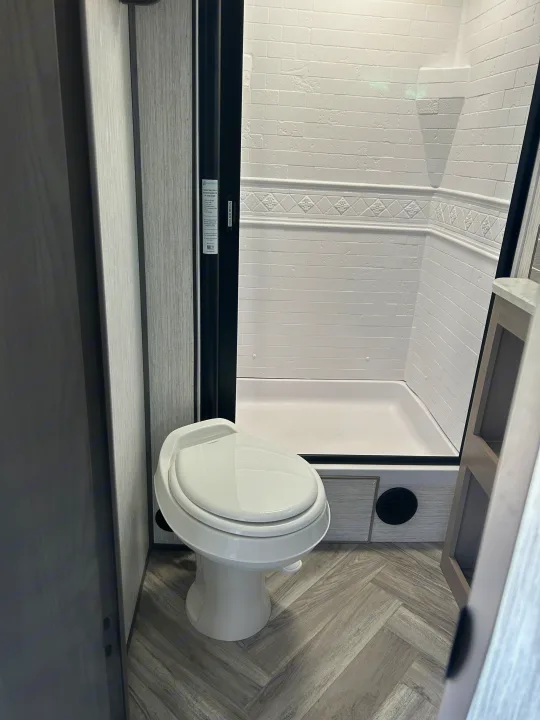 Interior of an RV rental bathroom with a white toilet, spacious shower stall featuring textured white walls, and herringbone-patterned floor.. Shadow Trailers Shadow Cruiser 2020
