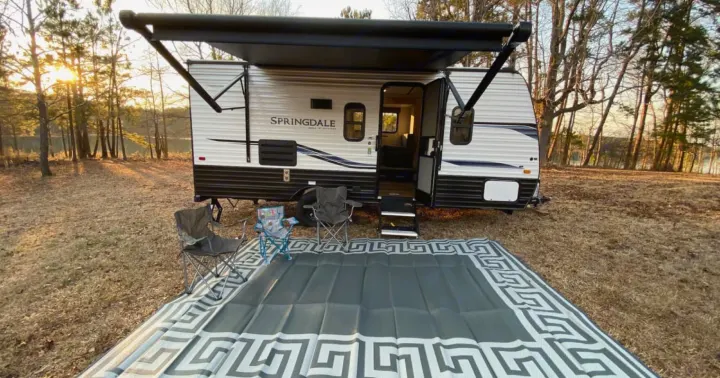 Peaceful RV rental setup by a lake at golden hour, featuring a travel trailer, an outdoor mat, and camping chairs.. Keystone RV Springdale 2022