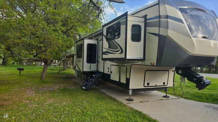 Enjoy your next getaway! A large Sandpiper fifth-wheel rv rental, with slide-outs extended, sits ready in a treed park.. Forest River Sandpiper 2021