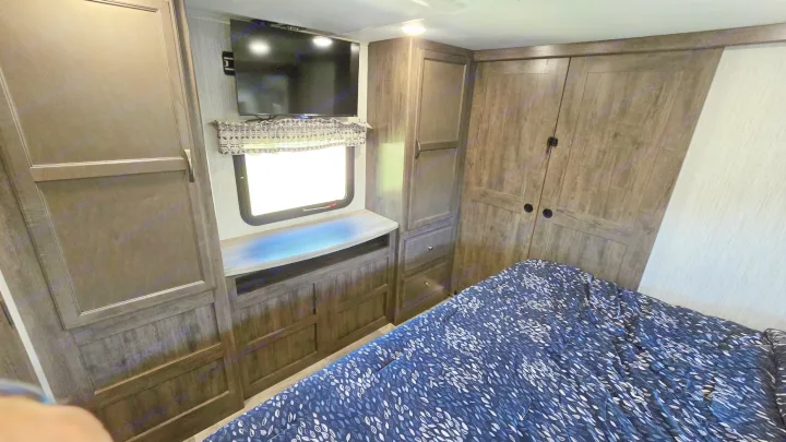 RV rental master bedroom with dark wood storage, a wall-mounted TV, and a patterned blue and white bed.. Forest River Sandpiper 2021