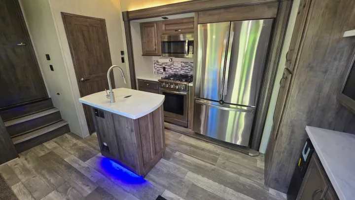 Modern RV rental kitchen featuring a stainless steel refrigerator, gas range, microwave, kitchen island with sink, and steps.. Forest River Sandpiper 2021