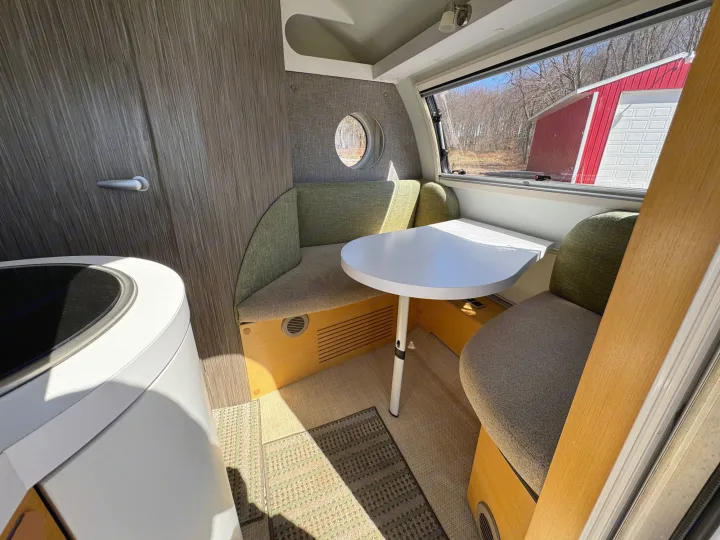 Cozy RV rental interior: round white table, green & gray banquette seating, with a view of a red barn and trees.. nuCamp T@B 2018