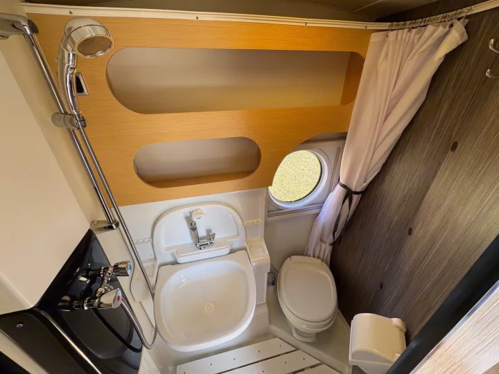 Inside an RV rental: a compact bathroom with shower, fold-down sink, toilet, shelves, and a porthole window.. nuCamp T@B 2018