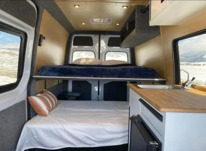 Cozy rv rental van interior featuring bunk beds, a compact kitchen, and windows showing a snowy landscape.. Vanworks Base Camp 2021