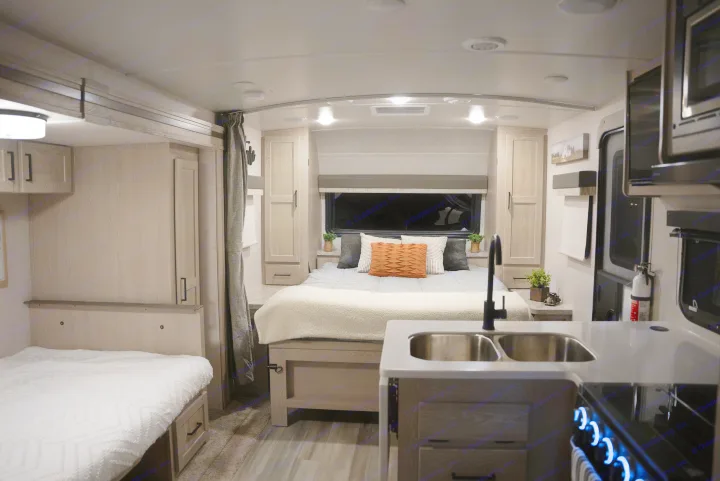 Inside a modern RV rental: a cozy bedroom with light wood cabinetry and a functional kitchen with a sink and lighted stovetop.. Forest River Flagstaff Micro Lite 2022