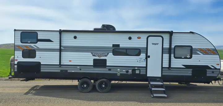 White & grey Wildwood travel trailer RV rental, a spacious recreational vehicle with black accents, wheels, and entry steps.. Forest River Wildwood X-Lite 2022