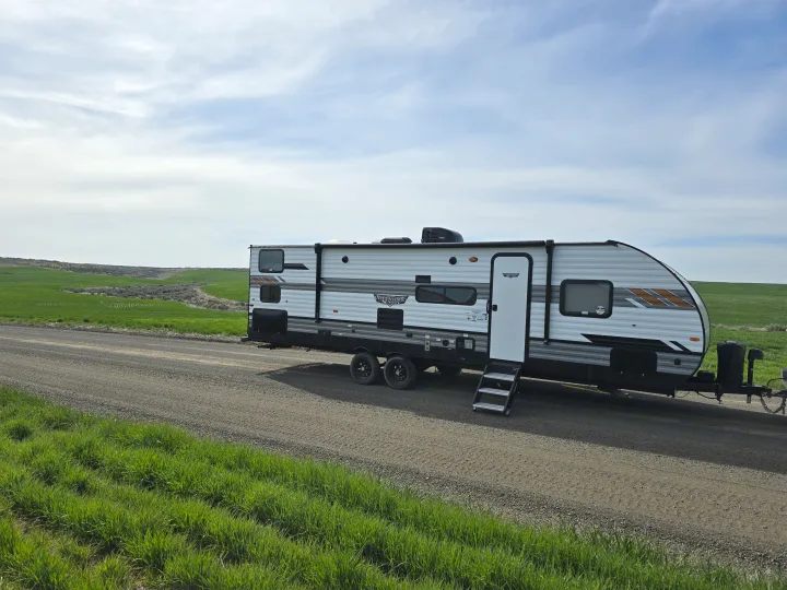 Modern travel trailer RV rental on a dirt road, ready for adventure in lush green fields under a cloudy sky.. Forest River Wildwood X-Lite 2022