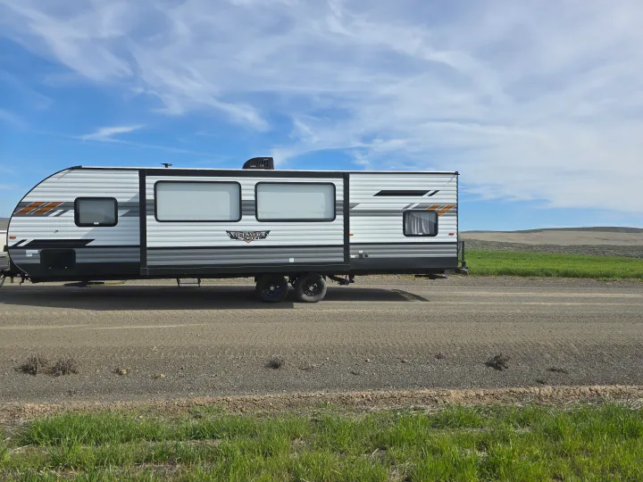 Your next RV rental adventure awaits! A modern travel trailer is parked on a gravel road by green fields & hills.. Forest River Wildwood X-Lite 2022