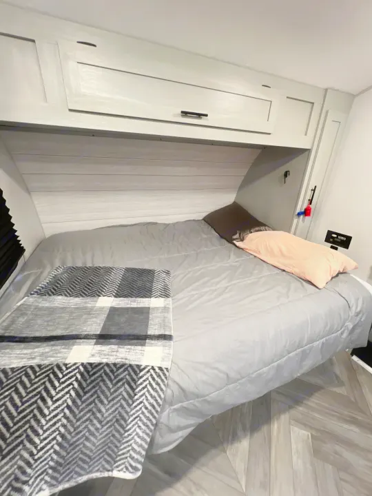 Cozy RV rental bedroom with a grey bed, plaid blanket, pillows, overhead cabinets, and wood-patterned floor.. Forest River Wildwood 2022